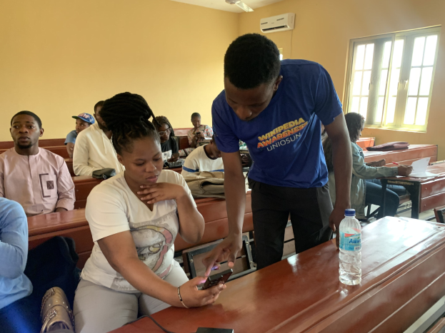 Eine Klassenszene mit Schülern an Tischen sitzend, ein Mann mit einem Mobiltelefon in der Hand stehend, Flaschen und andere Gegenstände auf den Tischen, eine Klimaanlage an der Wand und ein Fenster im Hintergrund.