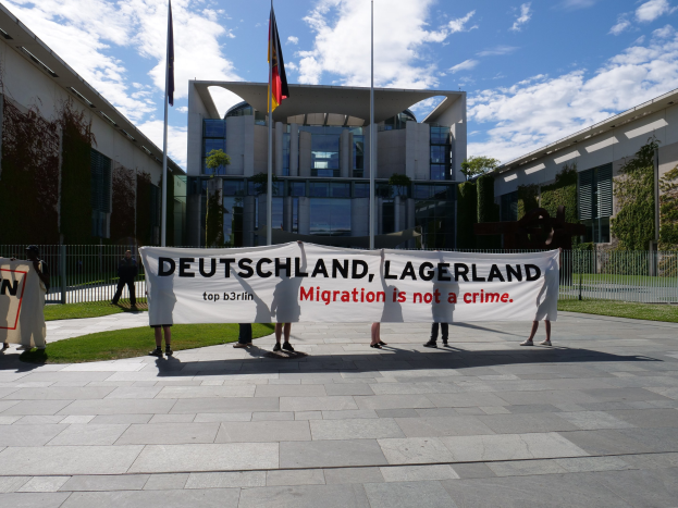 Gruppe von Menschen mit einem Transparent 'Deutschland, Lagerland Migration ist kein Verbrechen' vor einem Gebäude mit Fenstern, umgeben von Gras, einem Metallzaun, Pflanzen, Bäumen und Fahnen auf Stangen unter einem bewölkten Himmel.