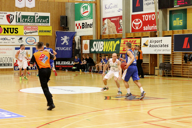 Ein Basketballfeld mit Spielern im Vordergrund und einer hölzernen Wand mit Postern und Banner im Hintergrund.