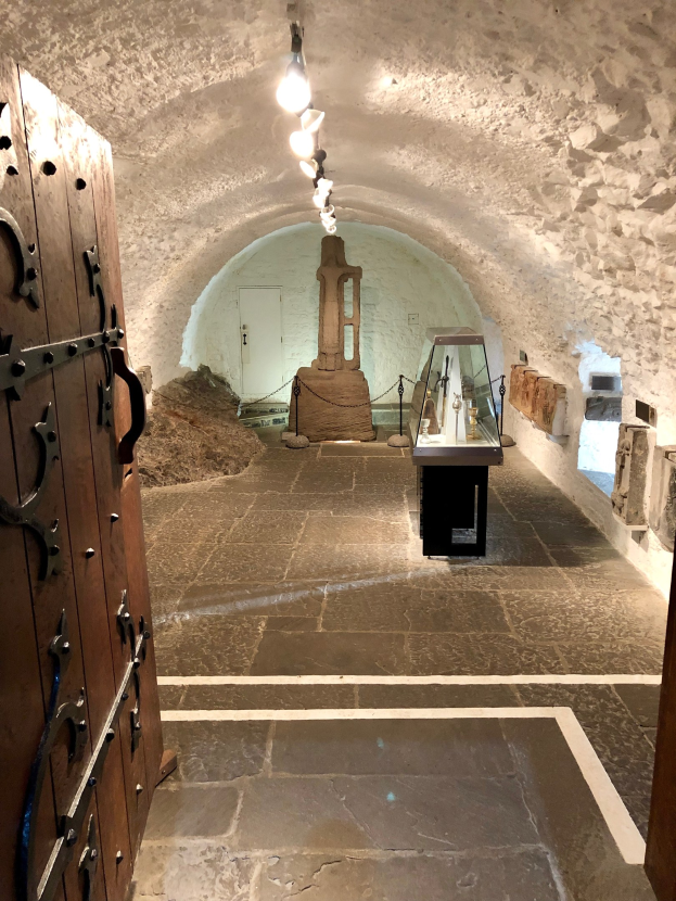 Eine hölzerne Tür auf der linken Seite, eine Steinwand im Hintergrund, Deckenlampen, die einen Tisch mit Gegenständen in der Mitte eines Raumes im Castello di San Giovanni, Italien, beleuchten.