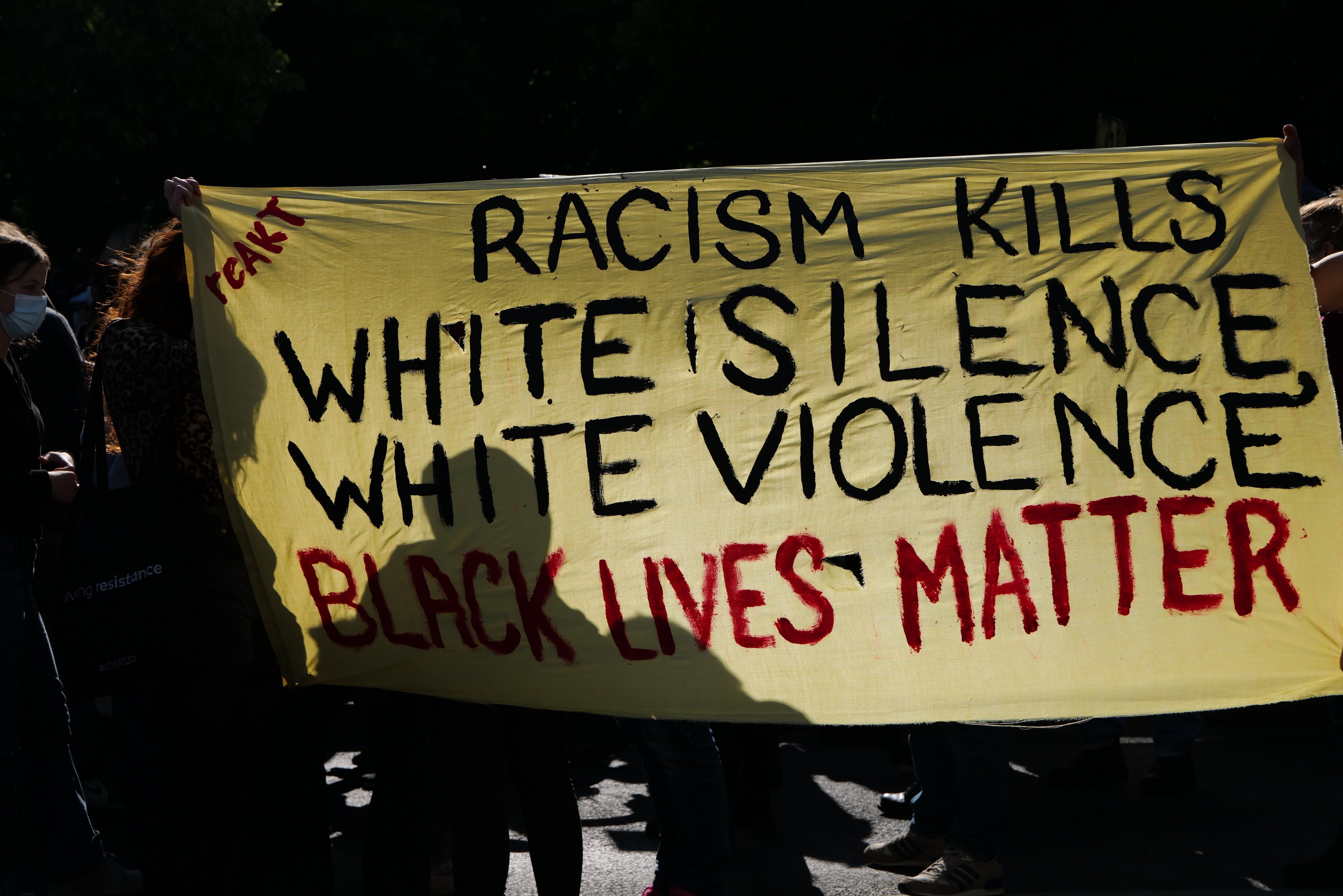 Eine Gruppe von maskierten Menschen steht nachts auf einer Straße und hält ein Transparent mit der Aufschrift "Rassismus tötet, weiße Stille, weiße Gewalt, Black Lives Matter."