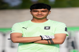 Ein junger Mann in einem grünen T-Shirt steht auf einem Fußballfeld mit verschränkten Armen und lächelt, mit einem unscharfen Hintergrund, der darauf hindeutet, dass er im Fokus der Aufnahme ist.