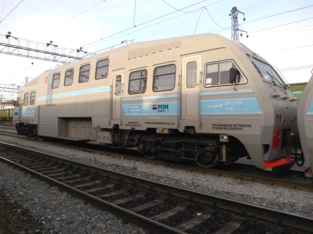 Eine diesel-elektrische Lokomotive fährt auf Schienen neben einem anderen Zug, mit elektrischen Masten und Kabeln im Hintergrund unter einem klaren blauen Himmel.