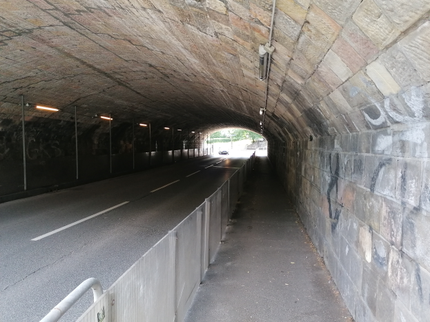 Ein Auto fährt durch einen beleuchteten Tunnel mit Graffiti an den Wänden, einem Geländer an der Seite und Bäumen im Hintergrund.