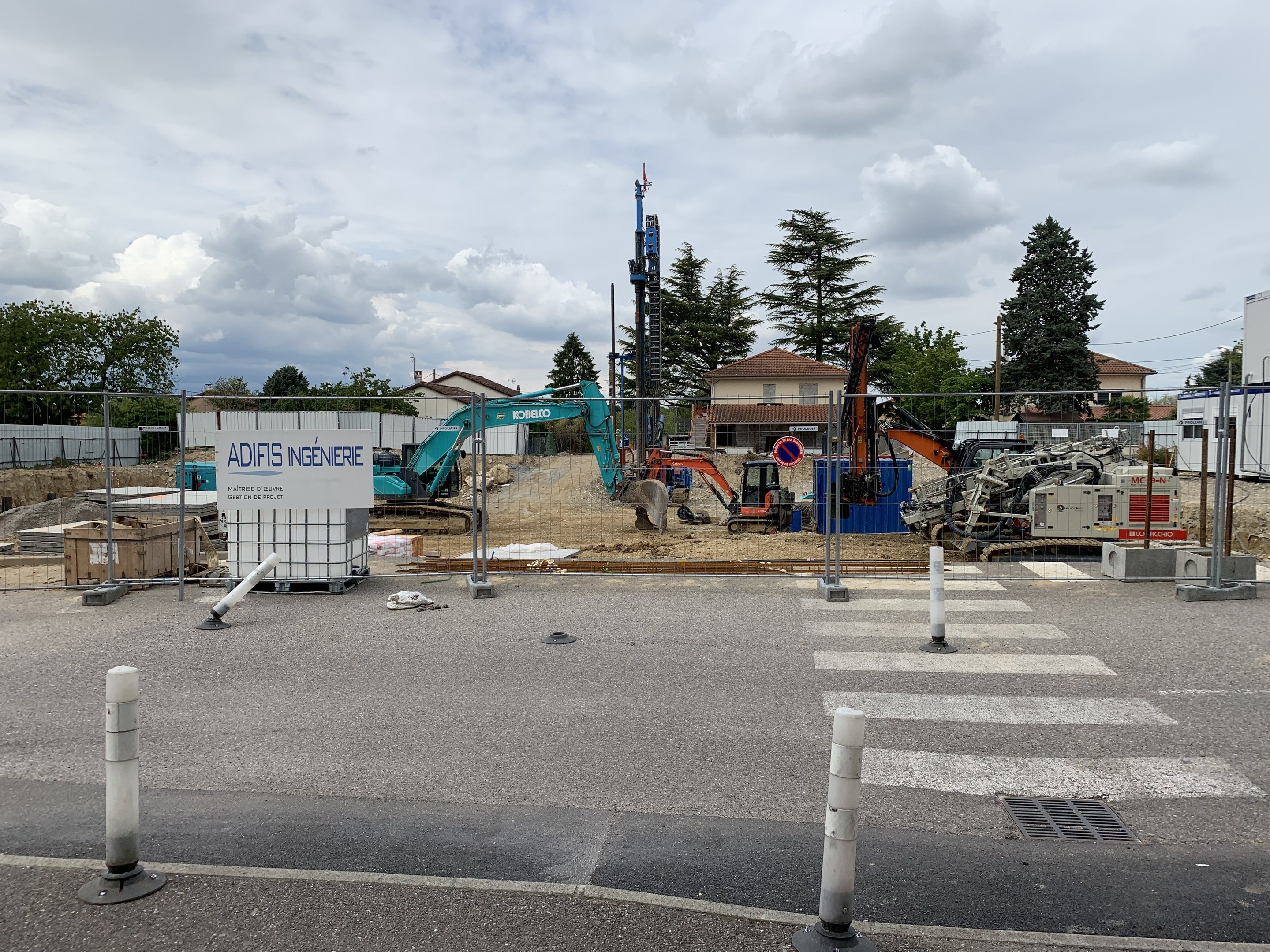 Baustelle mit verschiedenen Geräten, einschließlich Pfählen, einem Zaun, einer Tafel, Fahrzeugen, Bäumen, Häusern und einem bewölkten Himmel.