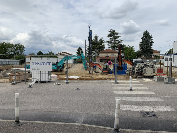Baustelle mit verschiedenen Geräten, einschließlich Pfählen, einem Zaun, einer Tafel, Fahrzeugen, Bäumen, Häusern und einem bewölkten Himmel.