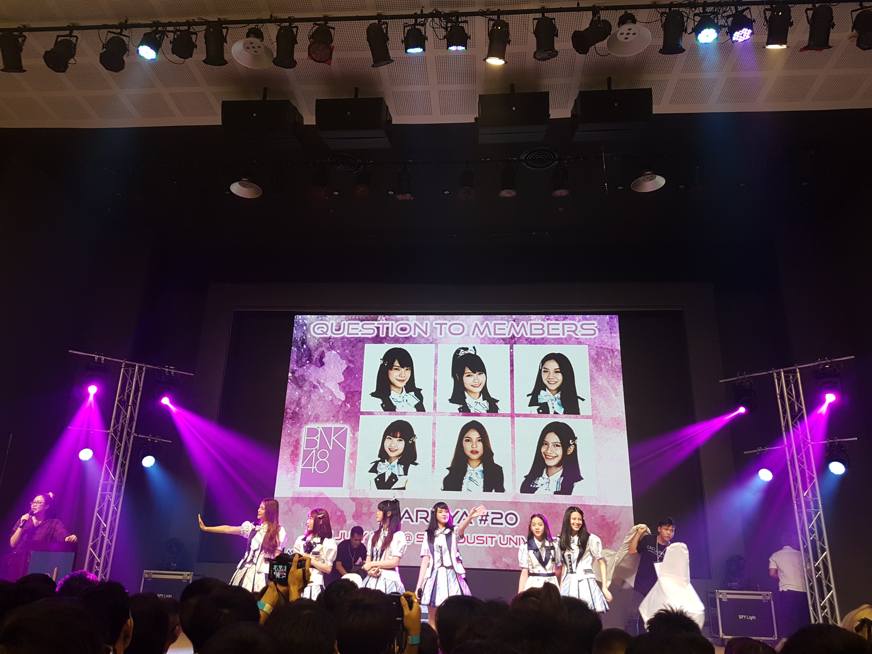 Eine Gruppe von Menschen performs auf einer hell erleuchteten Bühne mit Trägern, während ein Sänger ein Mikrofon auf der linken Seite hält und ein anderer ein Instrument spielt, während eine Menge darunter das Ereignis aufzeichnet; ein großer Bildschirm im Hintergrund zeigt Bilder der K-Pop-Gruppe BK48.