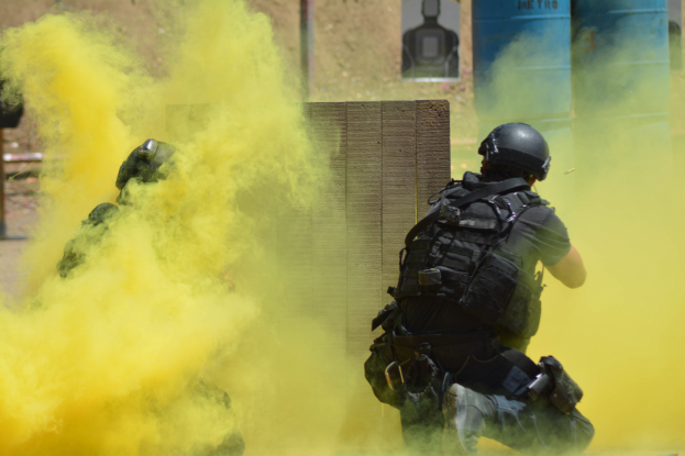 Zwei Polizisten in Einsatzausrüstung mit gelbem Rauch aus dem Mund, die neben einer Wand mit einer Tafel und anderen Gegenständen im Hintergrund stehen.