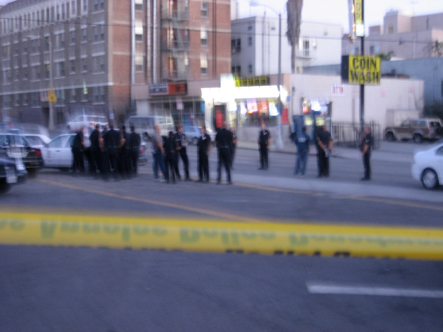 Eine Gruppe von Menschen, wahrscheinlich Offiziere, die auf einer Straße mit Autos, einem Band, Gebäuden mit Fenstern, einem Schild an einem Pfahl, Bannern, einem Zaun und Bäumen im Hintergrund stehen.