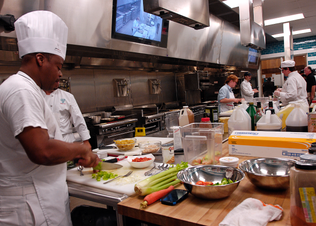 Eine Gruppe von Köchen in einer Küche, die auf einem Schneidbrett Lebensmittel zubereiten, mit einem Koch im Vordergrund in einer weißen Schürze und Mütze, der ein Messer hält, umgeben von verschiedenen Utensilien, Schüsseln, Gemüse, einem Mobiltelefon, Flaschen und anderen Gegenständen, mit einem Fernseher und Deckenleuchten im Hintergrund.