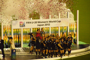 Eine Gruppe von Frauenfußballspielerinnen in schwarzen Uniformen feiert ihren Sieg auf einer Bühne, umgeben von einer Menge mit Kameras, mit einem dekorierten Hintergrund und grasbewachsenem Boden.