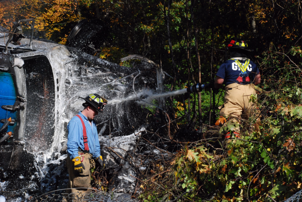 Zwei Feuerwehrleute in Helmen und Handschuhen, einer spritzt Wasser aus einer Rohrleitung auf ein umgestürztes Auto im Wald, mit Bäumen im Hintergrund und einem Maschendrahtzaun unten.