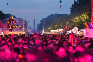 Eine große Menschenmenge auf einem nächtlichen Musikfestival, beleuchtet von bunten Lichtern und Ballons, mit Zelten, Bannern, Fahnen und anderen Gegenständen, die herumliegen, Bäumen, einem Turm und einem sternenklaren Himmel im Hintergrund.
