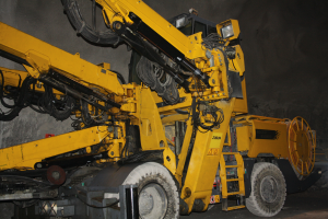 Ein großer gelber Bohrkrang mit langem Arm und Eimeranbau ist in der Mitte eines Tunnels positioniert, mit einer Wand im Hintergrund.