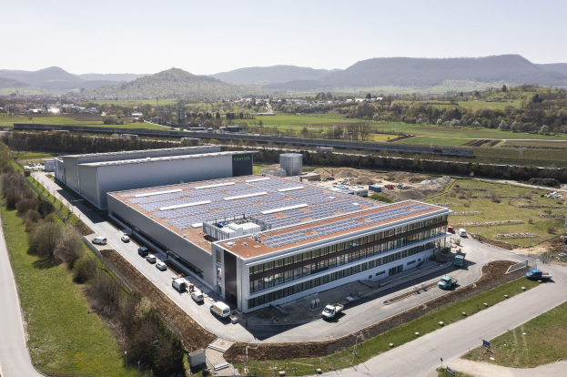 Luftaufnahme eines großen Gebäudes mit Solarmodulen auf dem Dach, umgeben von Fahrzeugen, Bäumen, Gras und einem klaren blauen Himmel in der Ferne.