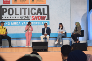 Eine Gruppe von Menschen sitzt auf einer Bühne mit einer Frau, die ein Mikrofon in der Mitte hält, einer Zuschauermenge im Vordergrund und einem Banner mit der Aufschrift "Politischer Roadshow 2019" im Hintergrund.