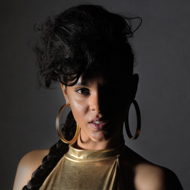 Eine Frau in einem goldenen Kleid und großen Kreolen steht selbstbewusst vor einer Wand, ihr glatter Bob-Haarschnitt und das natürliche Make-up unterstreichen ihren entschlossenen Gesichtsausdruck, während sie direkt in die Kamera blickt.