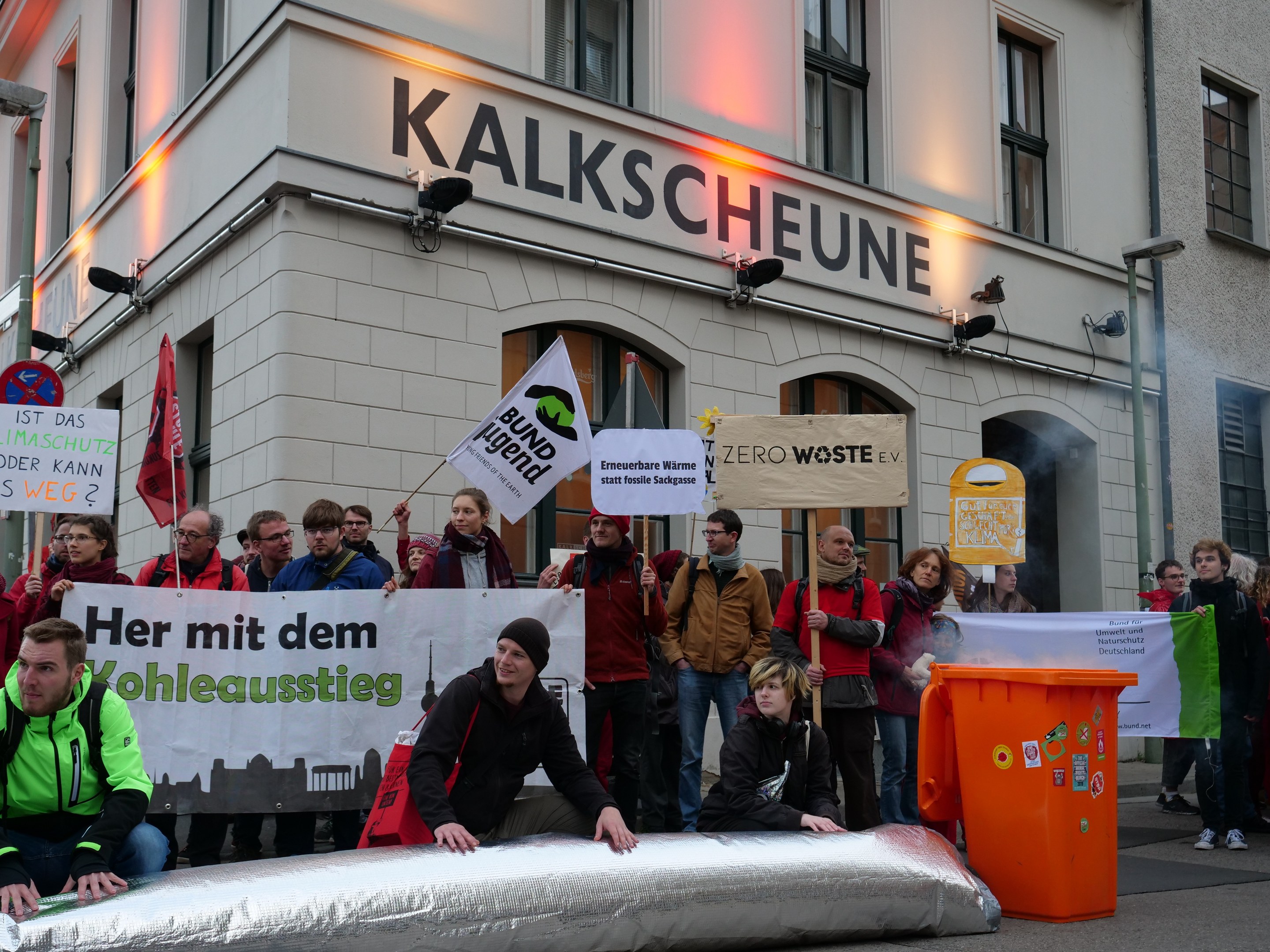 Gruppe von Menschen, die vor einem Gebäude in Deutschland protestieren und Schilder sowie Plakate halten, mit zwei Personen im Vordergrund und einem Müllcontainer auf der rechten Seite.