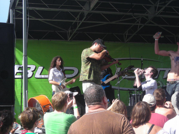 Eine Gruppe von Musikern, die verschiedene Instrumente auf einer Bühne spielen, mit einem großen schwarzen Lautsprecher links und einem grünen Banner im Hintergrund.