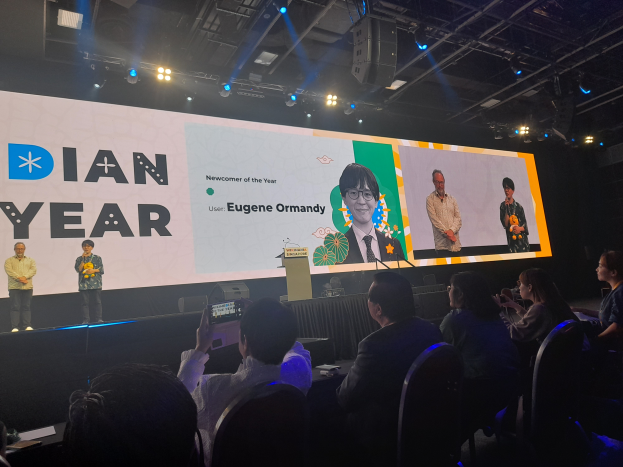 Eine Gruppe von Menschen sitzt auf Stühlen und schaut auf eine Bühne mit drei Personen, einem Podium mit Mikrofon, einem Tisch mit verschiedenen Gegenständen und einem Bildschirm mit Bildern und Text, unter einer Decke mit Lampen und Metallstangen, während eines Ereignisses, bei dem Eugene Ormandy, Gründer und CEO von Dian Year, anwesend ist.