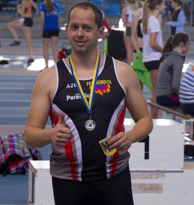 Ein Mann in einem schwarzen Jersey und einer Medaille steht, während einige Menschen im Hintergrund trainieren.