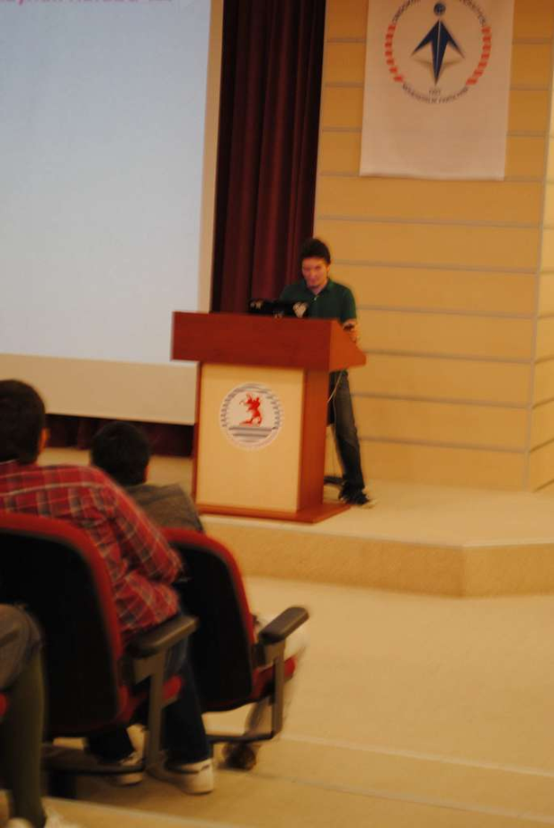 Eine Person steht an einem Podium in der Mitte, spricht, mit mehreren sitzenden Individuen links, einem Boden unten und Brettern und einem Vorhang links.