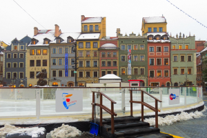 Eislaufbahn in einem Stadtplatz umgeben von Geb├Ąuden, Treppen, Gel├Ąndern, Informationsschildern, schneebedecktem Boden, Laternenmasten, B├Ąumen und einem Himmel im Hintergrund.