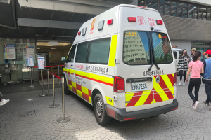 Ein Rettungswagen steht vor einem Gebäude, vor dem Menschen rechts entlanggehen, eine Person sitzt im Hintergrund auf einem Stuhl und Schilder mit der Aufschrift "Ambulance Service in Hong Kong" sind links zu sehen.