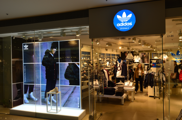 Ein Adidas-Laden in einem Einkaufszentrum mit Schaufensterpuppen, Kleidung an Bügeln, Schuhen auf Ständern und einem beleuchteten Adidas Originals-Logo-Schild.