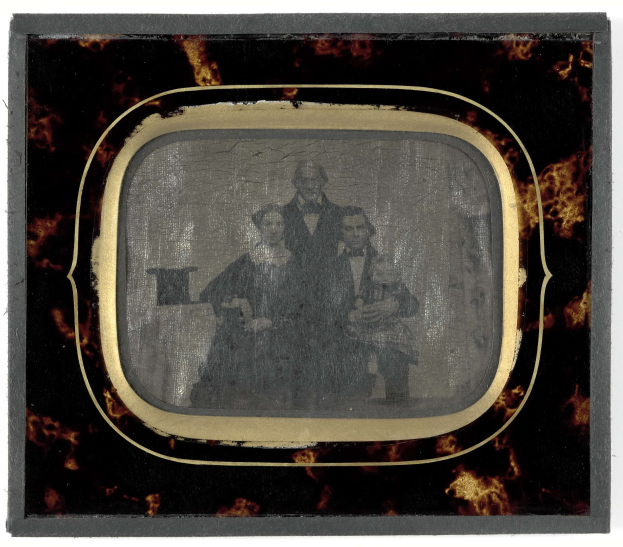 Schwarze-und-weißes Foto eines Mannes in Anzug und Krawatte und einer Frau in einem unbekannten Outfit, die vor einem Fernseher mit ernsten Gesichtern stehen.