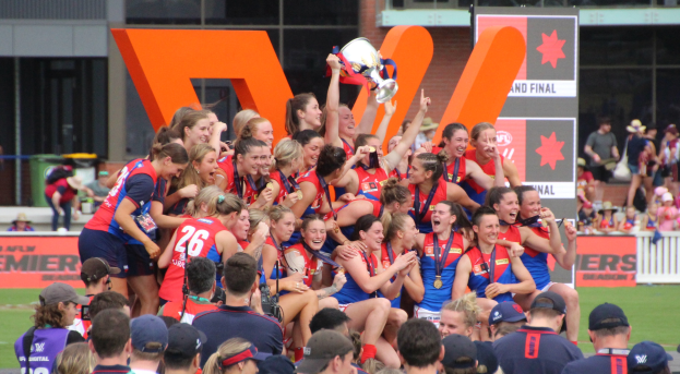 Eine Gruppe von Frauen-AFL-Spielerinnen in Sportuniformen feiert mit einem Pokal vor einem Publikum, einige halten ihn, mit Hintergrund-Elementen wie Schildern, Geländern, Menschen, einem Glasfenster-Gebäude und Gras.