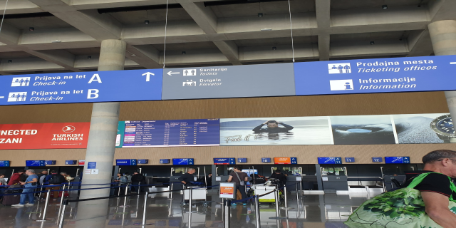 Menschen in einem Flughafen-Terminal in der Nähe eines "Check-in"-Schilds, einige halten Koffer, mit Pfosten, Bändern, Informationsschildern, Säulen und Deckenleuchten.