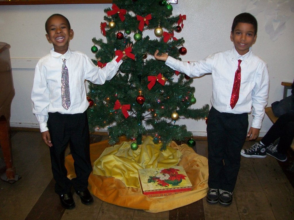 Ein geschmückter Weihnachtsbaum steht in einem Raum mit einer Kiste und anderen Gegenständen auf dem Boden, einem hölzernen Tisch auf der linken Seite, einem Stuhl auf der rechten Seite und einer weißen Wand im Hintergrund mit Gegenständen daran befestigt; zwei Jungen in weißen Hemden lächeln, während sie den Baum halten.