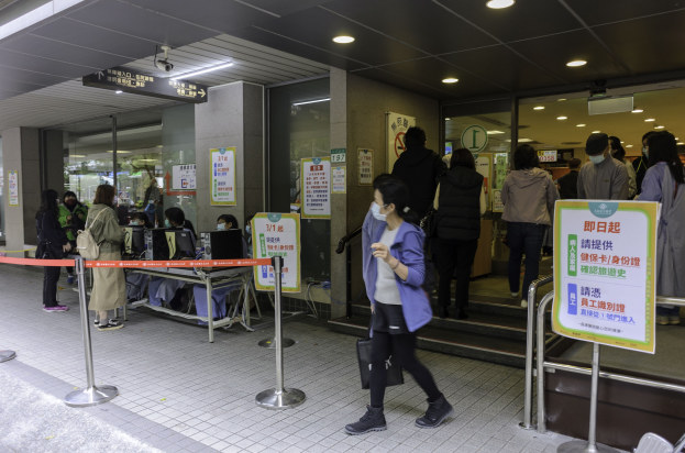 Eine Frau mit Gesichtsmaske geht an einer Warteschlange vorbei an einem Bahnhof, mit Metallstangen, Schildern, Papieren an einer Wand, einer Überwachungskamera und Deckenleuchten.
