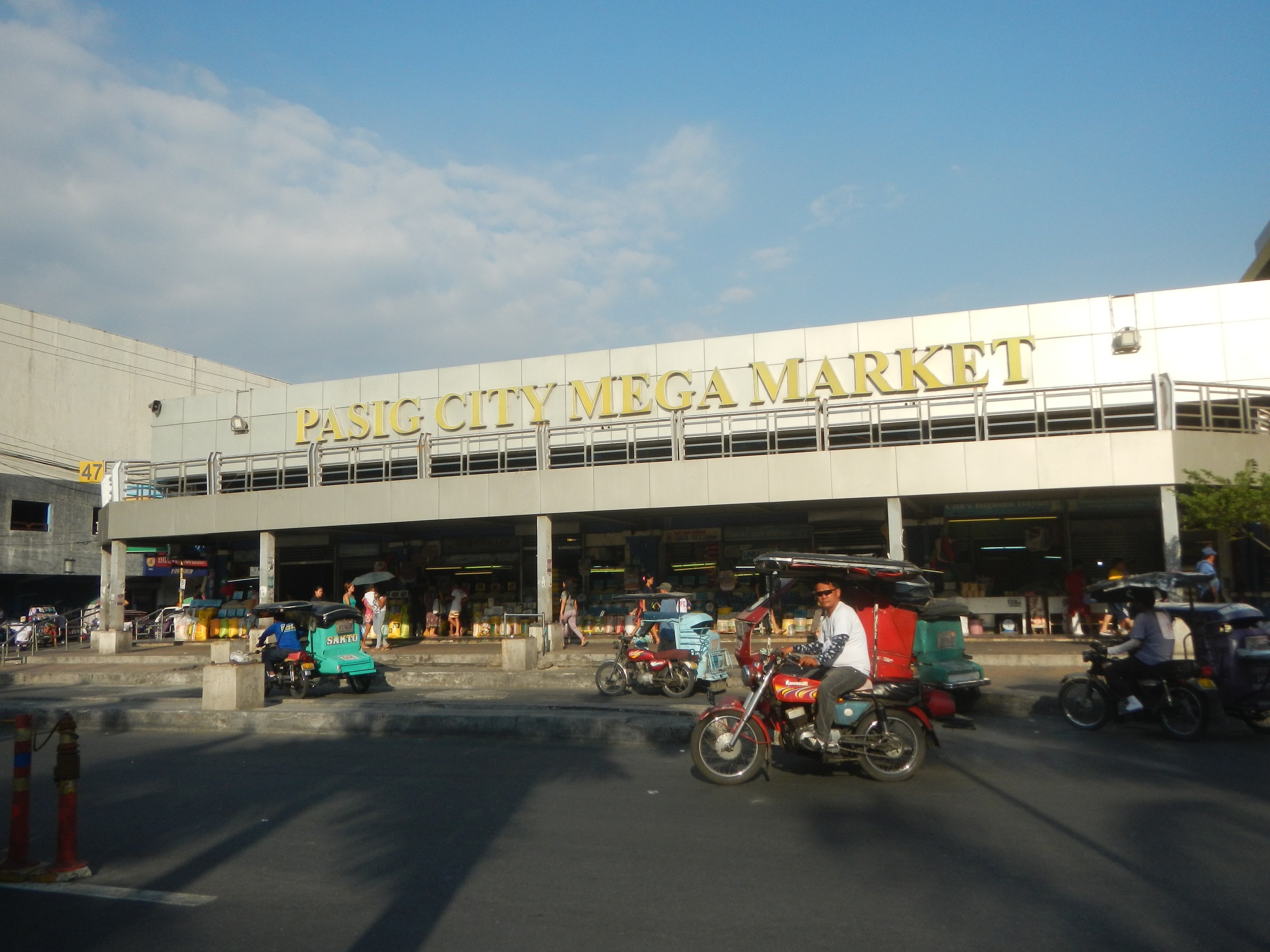 Eine belebte Straßenszene am Pasig City Mega Market mit Fahrzeugen und Fußgängern, einem Gebäude mit Text im Hintergrund und einem bewölkten Himmel.