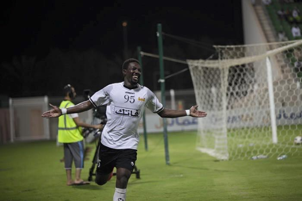 Ein Mann in weißem Hemd und schwarzen Shorts feiert auf einem Fußballfeld mit ausgebreiteten Armen, umgeben von Zuschauern, in der Nähe eines Torpfostens mit Netz, während andere auf Treppen, Pfosten und einer Wand unter einem dunklen Himmel sitzen.