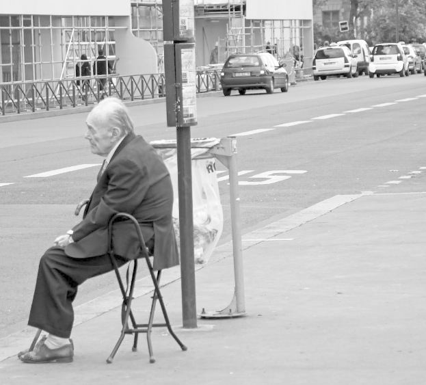 Ein älterer Mann in einem Anzug sitzt in einem Stuhl mit einer Straße und Autos im Hintergrund.