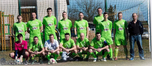 Eine Gruppe von Männern in grünen T-Shirts steht und hockt auf einem Fußballfeld mit einem Ball davor, ein Torpfosten rechts und ein Hintergrund aus Bäumen, Gebäuden, Pfählen, Fahrzeugen und Himmel, mit Text in der linken unteren Ecke, der die Zahlen von 1 bis 72 auflistet.