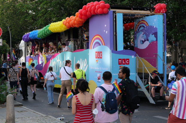 Eine Gruppe von Menschen, die neben einem Lastwagen mit bunten Luftballons die Straße entlanggehen, mit Schildern an Pfosten an der Straße und Bäumen und Gebäuden im Hintergrund, was auf eine Pride-Parade in Paris hindeutet.