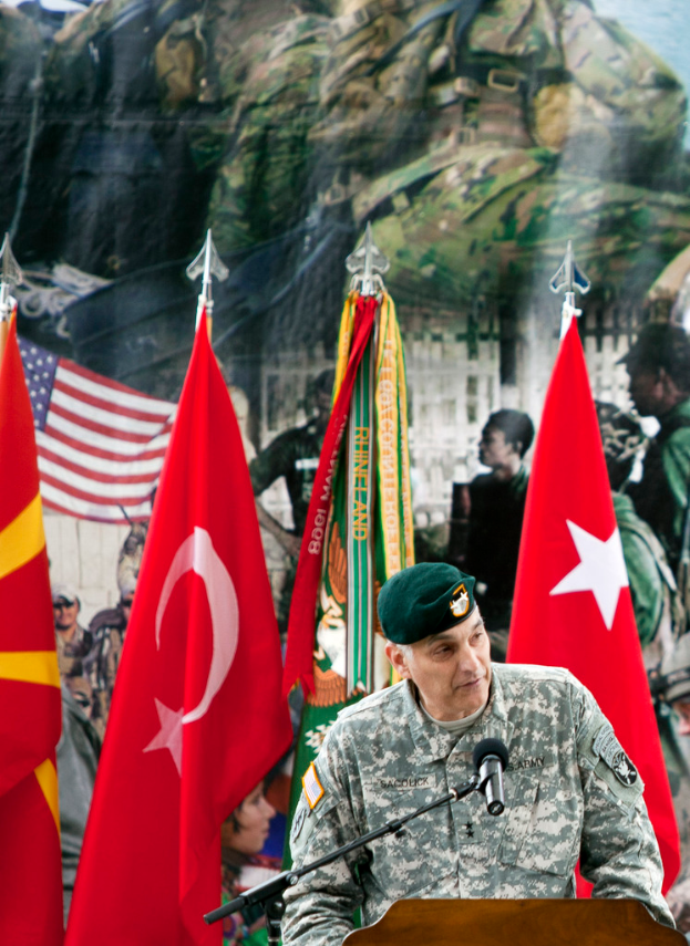 Ein Soldat steht auf einem Podium mit einem Mikrofon, mit verschiedenen Flaggen und einem Plakat mit Militärpersonal im Hintergrund.