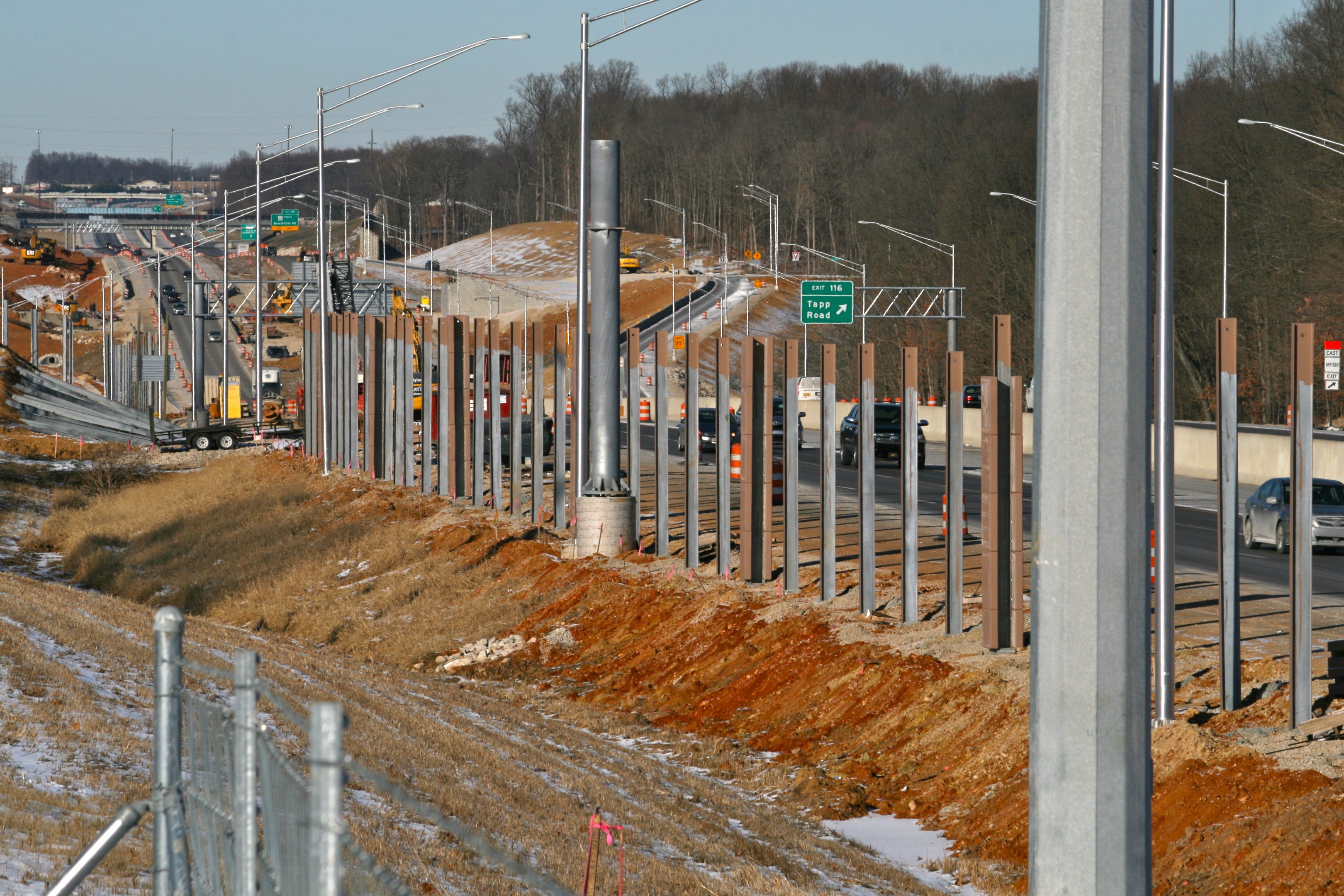 Baustelle mit Fahrzeugen, Polen, Lichtern, Beschilderung, einem Zaun, schneebedecktem Gras, Bäumen und einem Himmel im Hintergrund.