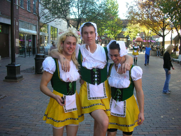 Drei Männer in weißen und gelben Kostümen stehen lachend vor einer Straßenszene mit Menschen, Gebäuden, Bäumen, einer Bank, einem Mülleimer und einem Geländer.