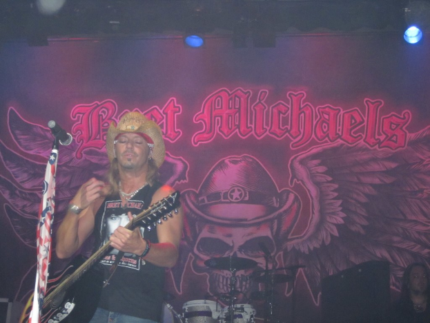 Eine Musik-Konzert-Szene mit einem beleuchteten Banner im Hintergrund, einer Person, die Gitarre spielt und in ein Mikrofon singt, Drogen in der Mitte unten und einer weiteren Person, die in der rechten unteren Ecke sitzt.