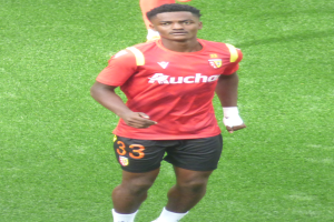 Emmanuel Adebayor, ein Mann in rotem T-Shirt und schwarzen Shorts, läuft auf einem Fußballfeld mit zwei Personen im Hintergrund.