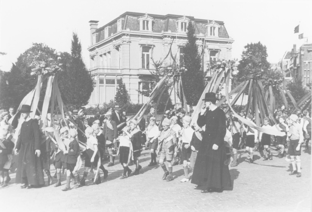 Schwarzes und weißes Bild einer Gruppe von Menschen, die eine Straße entlanggehen, einige halten Fahnen, mit Hüten, Maibaum, Bäumen, Gebäuden und Himmel.