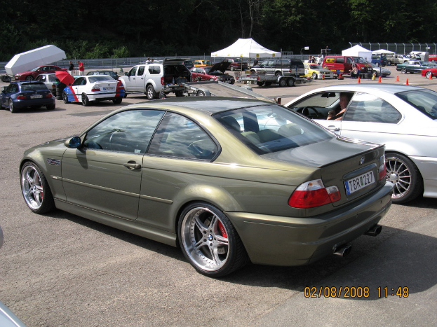 Eine Au├čenaufnahme mit mehreren Fahrzeugen auf einem Parkplatz, einem weißen Zelt in der Ferne, zahlreichen B├Ąumen, einigen Menschen, die stehen, einem roten Schirm und zwei Autos im Vordergrund.