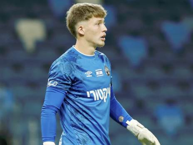 Ein junger Mann in einem blauen Torwarttrikot und weißen Handschuhen steht auf einem Fuballfeld während eines Spiels, mit einem unscharfen Hintergrund.