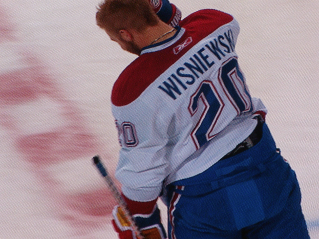 Ein Hockey-Spieler in einem "Wisniewski"-Trikot steht auf dem Eis, eine Hockey-Schläger haltend, mit einem konzentrierten und entschlossenen Gesichtsausdruck.