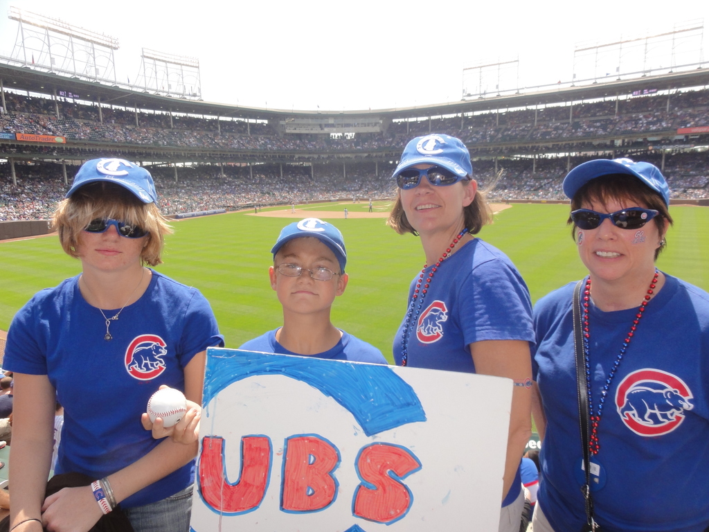 Drei Frauen mit Kappen und Brillen, ein Junge mit Brille und Kappe, der einen Ball und ein Schild hält, in einem Stadion mit Zuschauern und Feld im Hintergrund, unter einem Himmel mit einigen Lichtern.