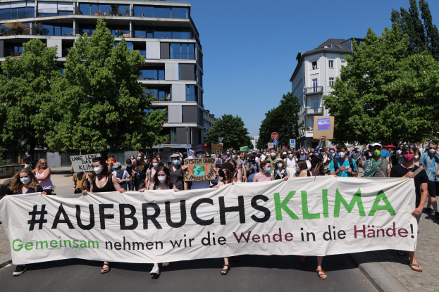 Eine Gruppe von Menschen mit Masken hält ein Banner mit der Aufschrift "Aufbruchsklima" vor einem Gebäude mit Bäumen und einem klaren Himmel.